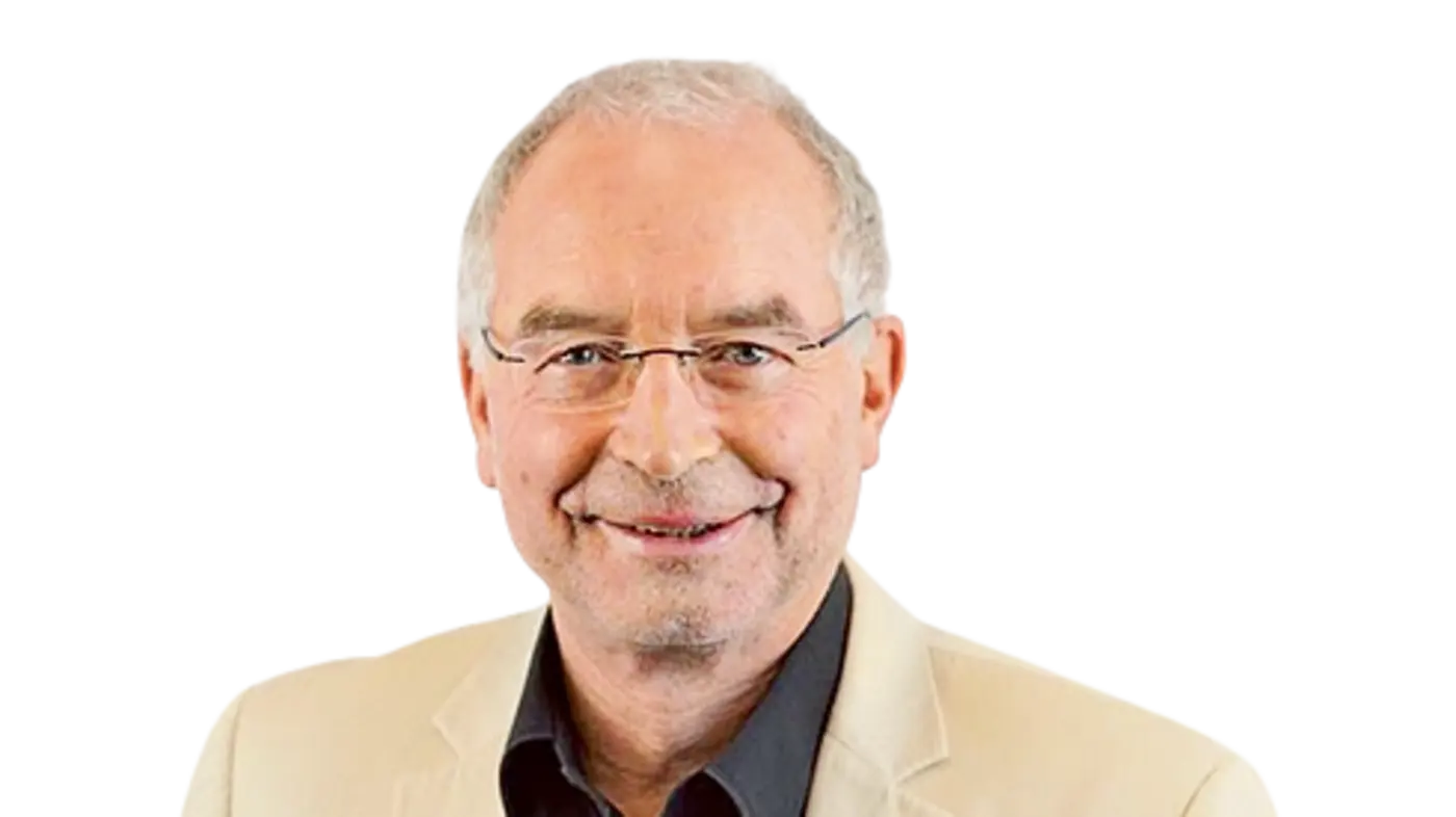 Porträt eines älteren Mannes mit grauen Haaren und Brille, der in die Kamera lächelt. Er trägt ein beigefarbenes Sakko über einem schwarzen Hemd. An seinem Revers ist ein Namensschild befestigt, das auf seine Funktion in einer medizinischen Einrichtung hinweist.