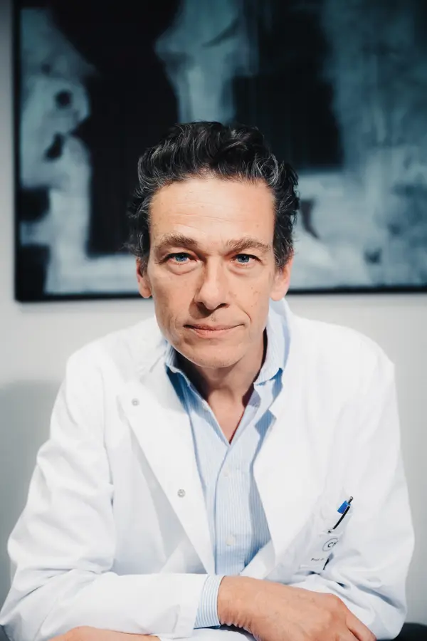 Ein Arzt im weißen Kittel blickt ernst in die Kamera, sitzt an einem Tisch; im Hintergrund ein unscharfes Bild an der Wand.