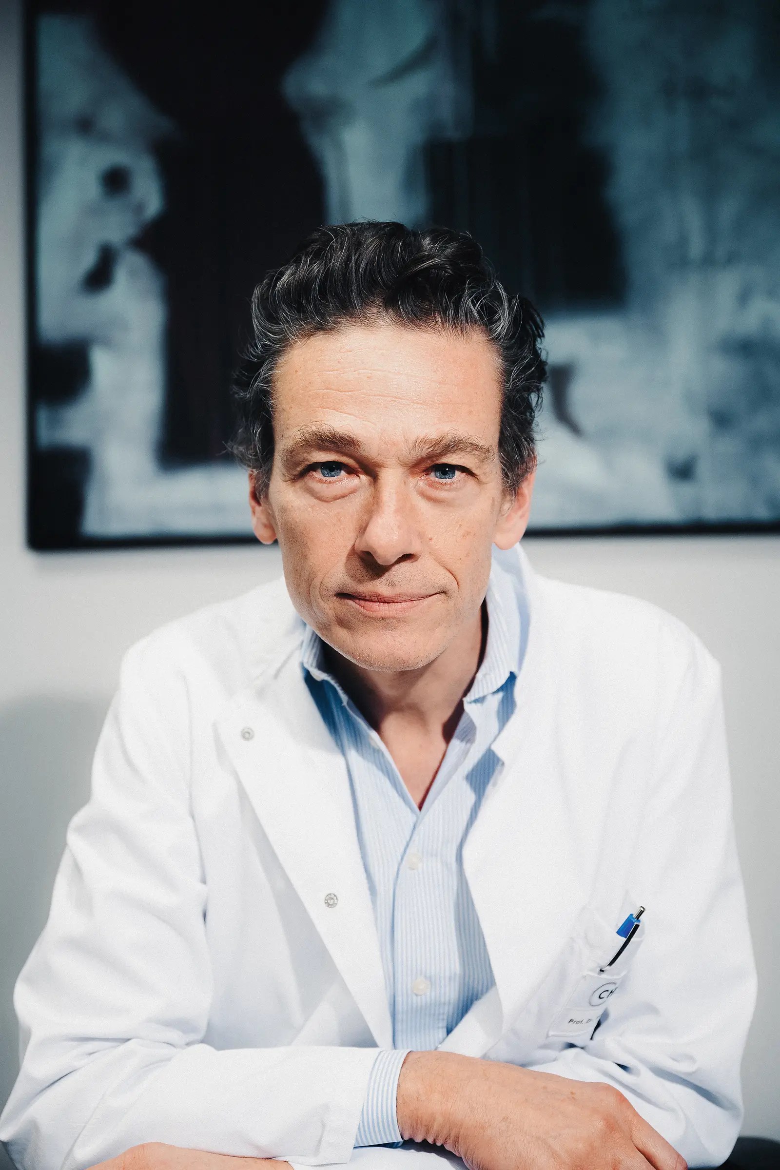 Ein Arzt im weißen Kittel blickt ernst in die Kamera, sitzt an einem Tisch; im Hintergrund ein unscharfes Bild an der Wand.