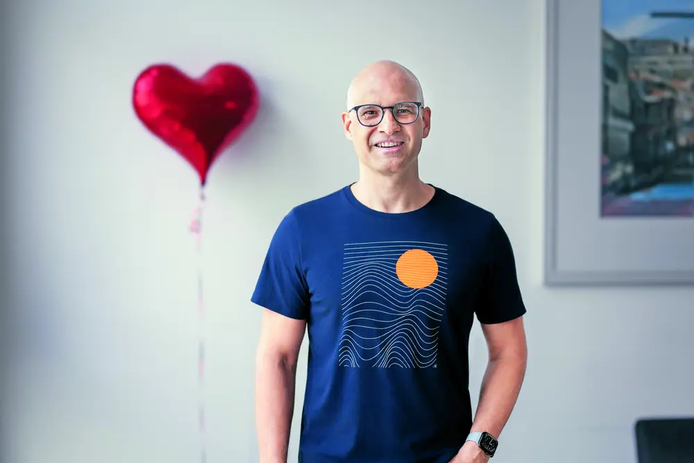 Das Bild zeigt einen Mann mit Glatze und Brille der in die Kameralächelt. Im Hintergrund ich ein roter Folienluftballon in der Form eines Herzes zu sehen. 