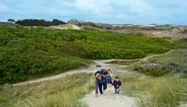 Eine Frau wandert mit einem Kind an der Hand und weiteren Begleitern eine Sanddüne hinauf, im Hintergrund ist eine grüne