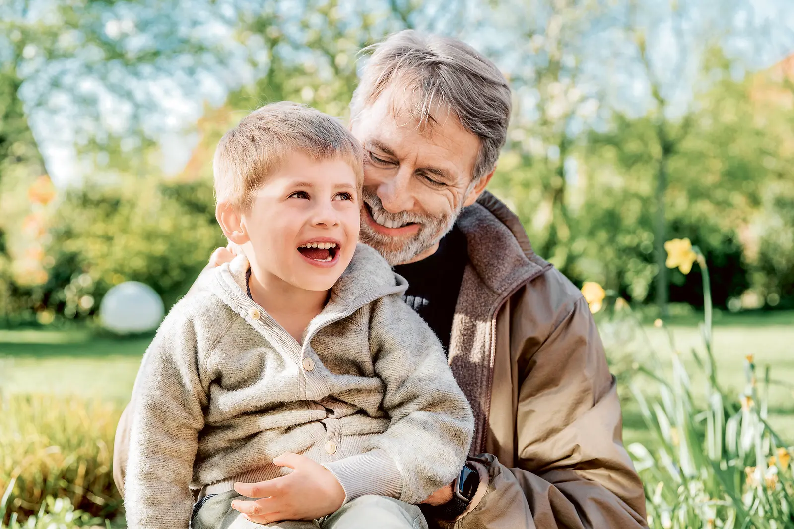 Ein älterer Mann mit grauen Haaren und Bart sitzt im Garten und hält einen kleinen Jungen auf dem Schoß. Beide lachen herzlich. Der Junge trägt eine beige-graue Strickjacke, der Mann eine braune Jacke über einem dunklen Pullover. Im Hintergrund sind grüne Wiese, Bäume und gelb blühende Narzissen zu sehen.