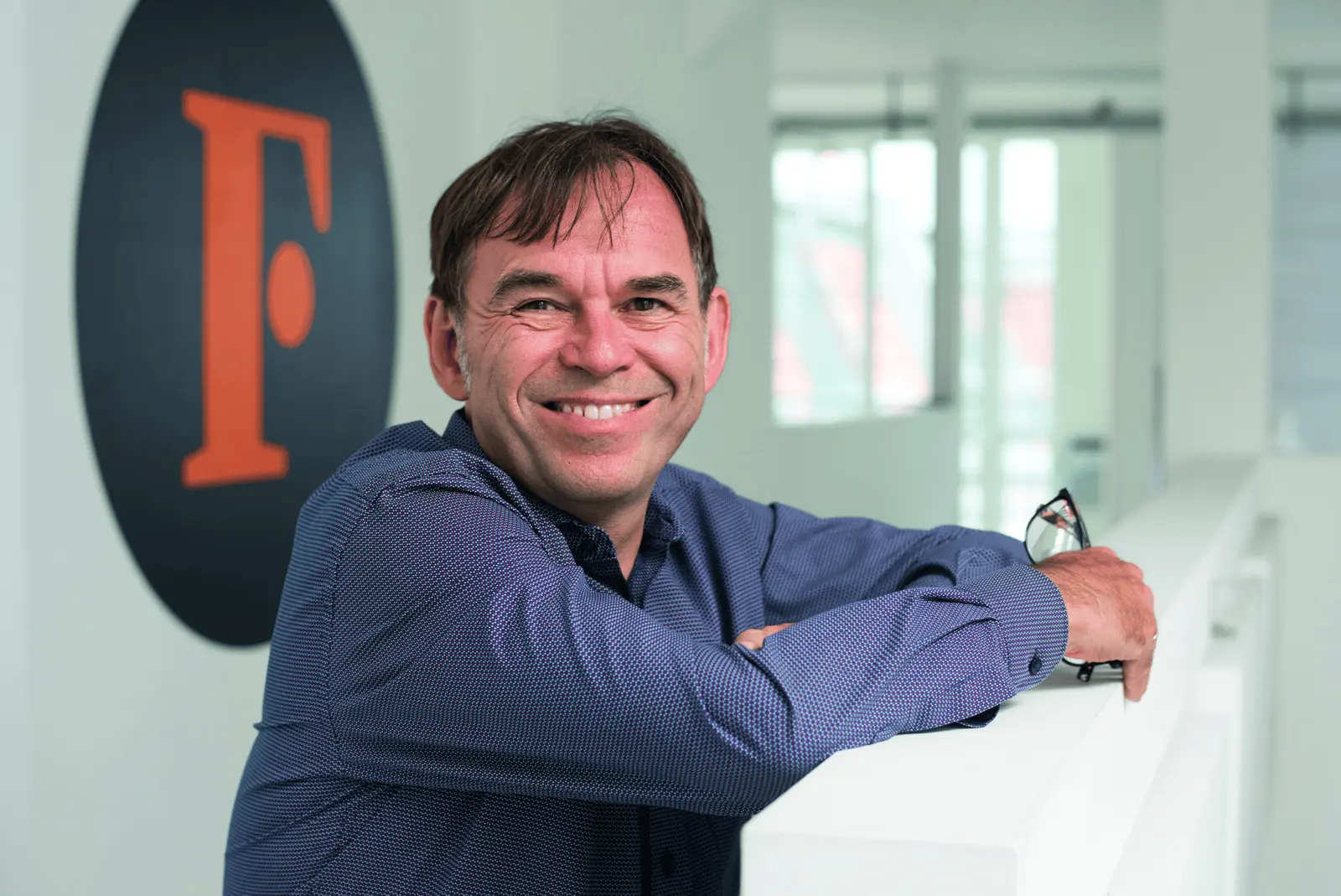 Porträt eines lächelnden Mannes in einem blauen Hemd, der sich an ein weißes Geländer lehnt und eine Brille in der Hand hält, neben einem Wandlogo mit einem orangen „F“.