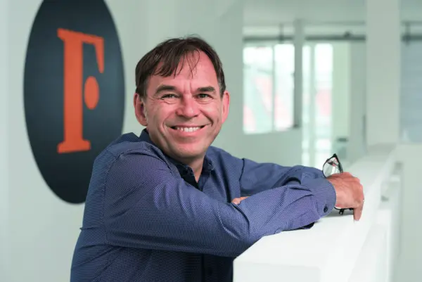 Porträt eines lächelnden Mannes in einem blauen Hemd, der sich an ein weißes Geländer lehnt und eine Brille in der Hand hält, neben einem Wandlogo mit einem orangen „F“.