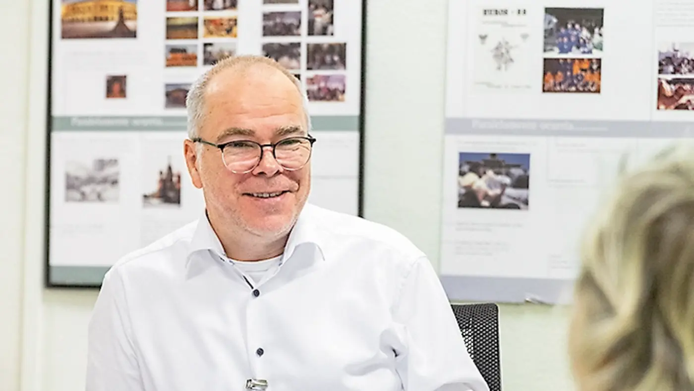 Ein Mann mit Brille und weißem Hemd sitzt lächelnd an einem Schreibtisch vor einem Laptop. Ihm gegenüber sitzt eine Frau, von der nur der Hinterkopf zu sehen ist. Im Hintergrund hängen Pinnwände mit Fotos und Dokumenten.