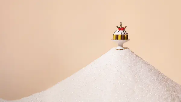 Ein kleiner Kuchen steht auf einem großen Berg aus Zucker vor hellem, minimalistischem Hintergrund.