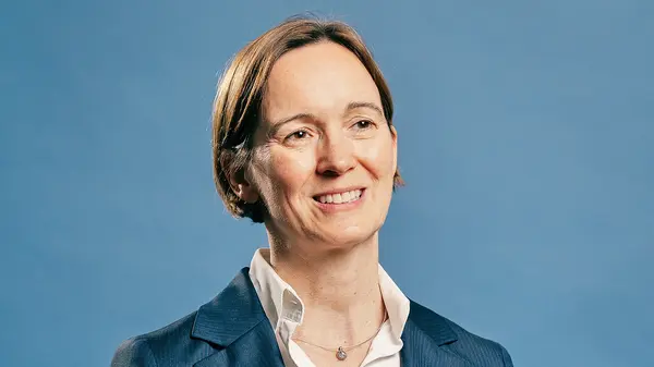 Porträt von Hilke Herchen vor blauem Hintergrund. Die lächelnde Juristin trägt einen dunklen Blazer über einer weißen Bluse und unterstreicht ihre Worte mit einer lebendigen Handgeste.