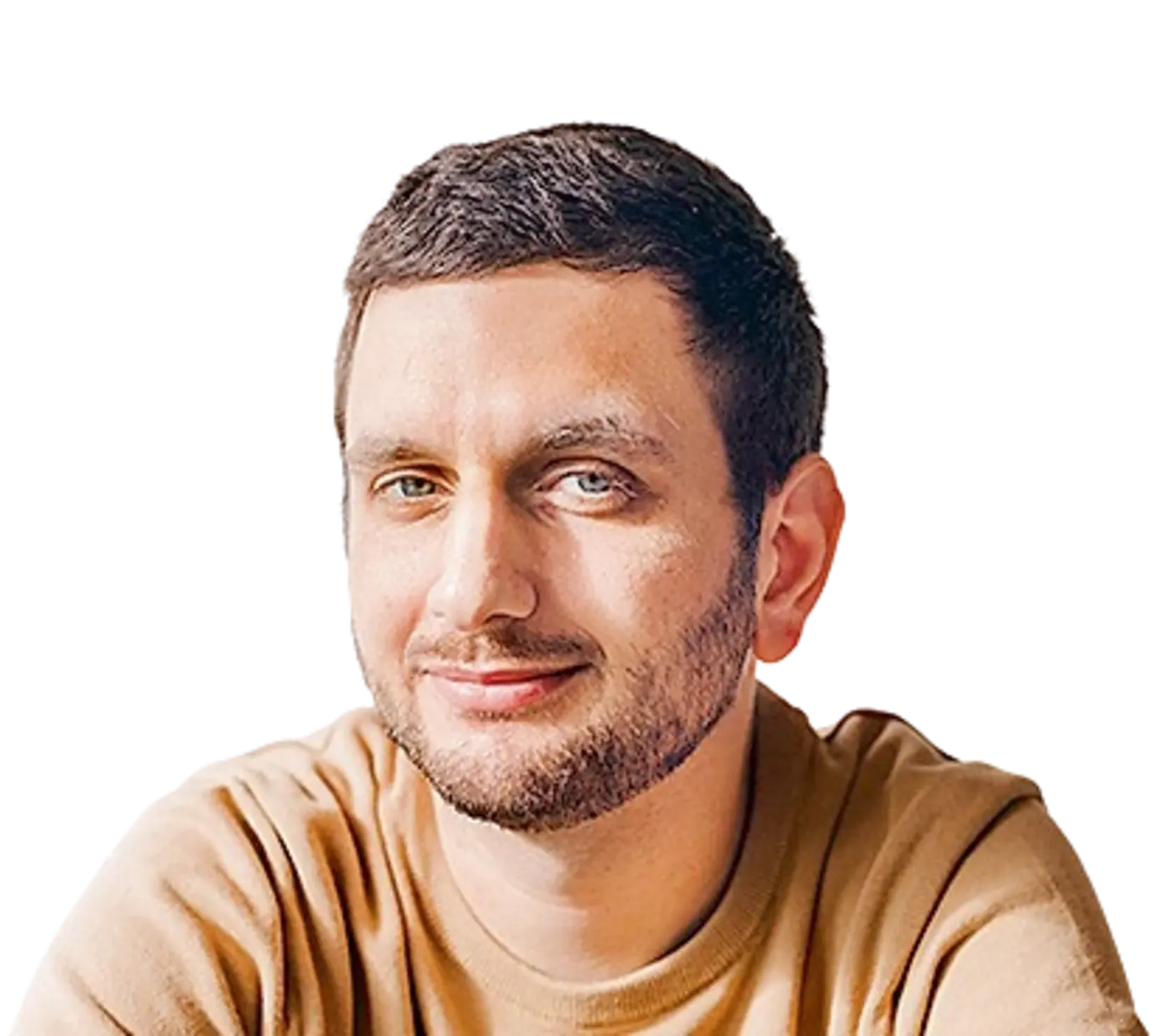 Das Bild zeigt einen Mann mit einem gepflegten Bart, der in einem beigefarbenen Pullover sitzt. Er hat ein leichtes Lächeln im Gesicht und schaut in die Kamera. Der Hintergrund ist schlicht und hell, und seine Hände sind auf einem Tisch abgelegt. Er wirkt freundlich und entspannt.