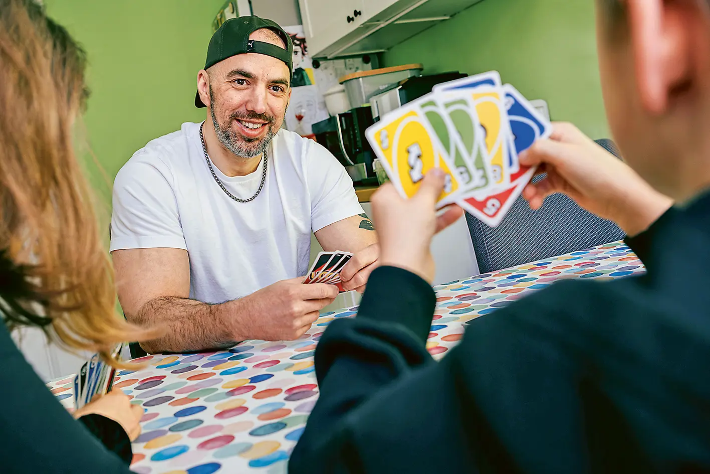 Ein Mann mit weißem T-Shirt, Kette und rückwärts getragener Basecap sitzt lächelnd an einem bunt gepunkteten Tisch und hält Spielkarten in der Hand. Ihm gegenüber sitzen zwei Kinder, deren Gesichter nicht zu sehen sind – eines davon hält auffällig bunte UNO-Karten. Im Hintergrund sind eine grüne Wand, Küchenschränke und Kinderzeichnungen zu erkennen.