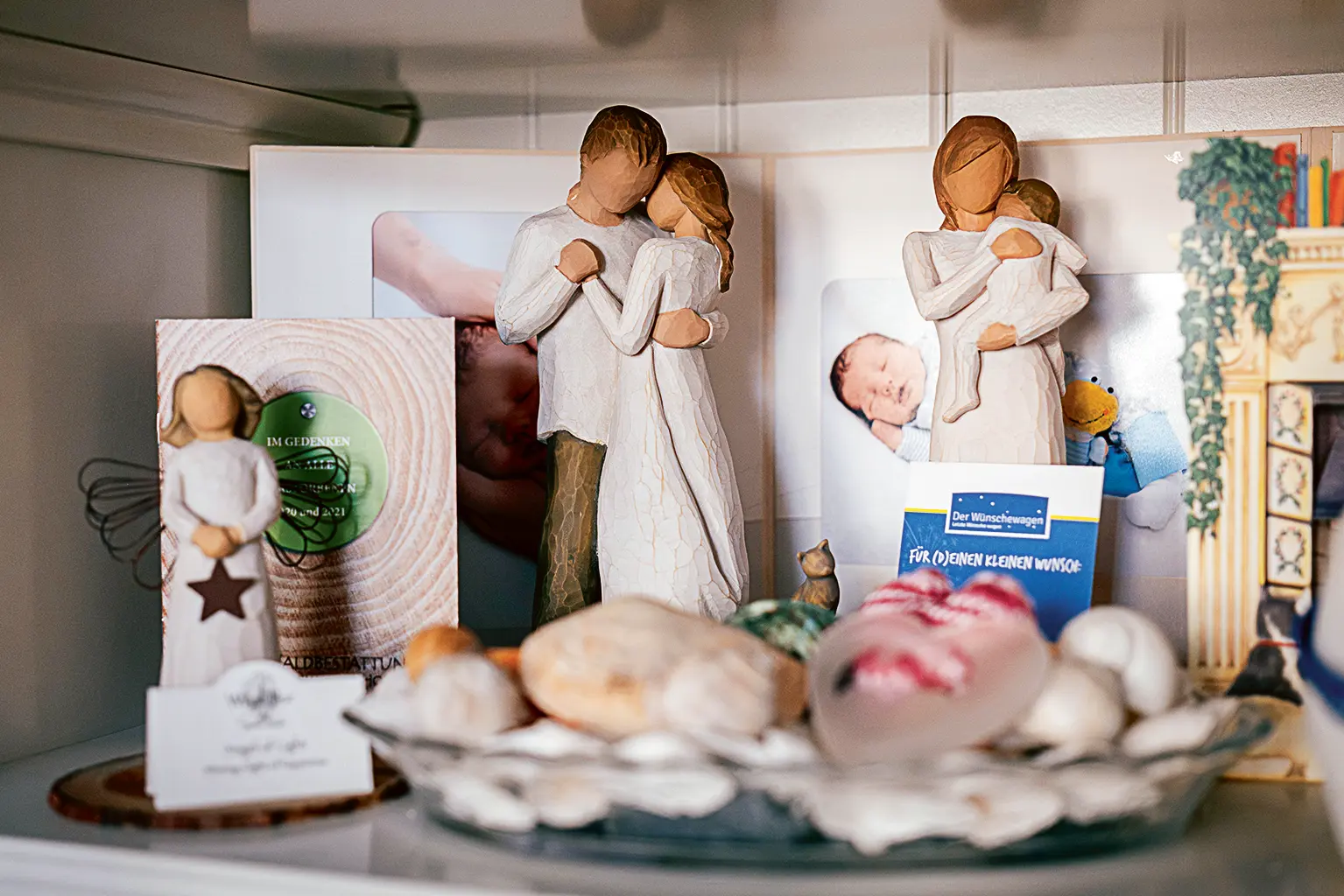Ein Regalbrett mit mehreren dekorativen Holzfiguren ohne Gesichtszüge, die innige Umarmungen darstellen – darunter Eltern mit Kind. Dazwischen stehen Erinnerungsstücke wie Muscheln, ein kleines Katzenfigürchen, eine Kerze in Herzform, Fotos eines Neugeborenen sowie Karten, darunter eine mit der Aufschrift „Der Wünschewagen – Für (d)einen kleinen Wunsch“ und eine Gedenkkarte mit der Aufschrift „Im Gedenken