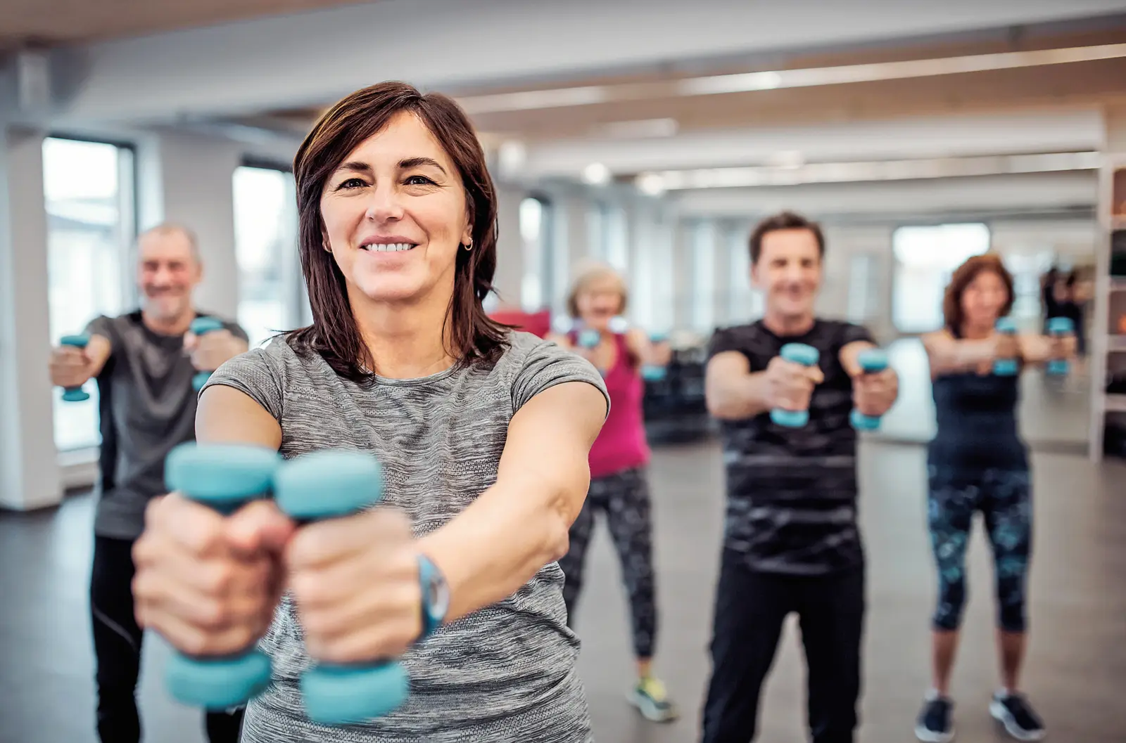 Eine Frau trainiert mit Hanteln in einem Fitnessraum; hinter ihr üben mehrere Männer und Frauen gemeinsam Kraftübungen.