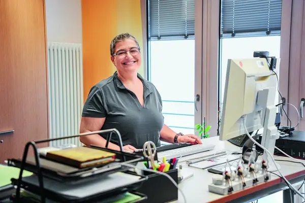 Eine Dame steht vor ihrem Arbeitsplatz und lächelt in die Kamera.