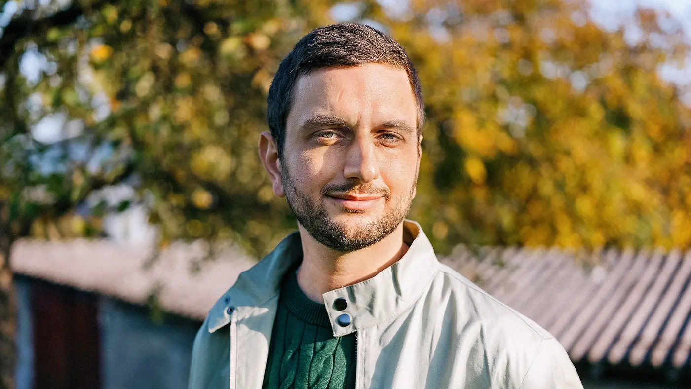 Ein Mann mit Bart steht im Freien, trägt eine helle Jacke und einen grünen Pullover; im Hintergrund herbstliche Bäume.