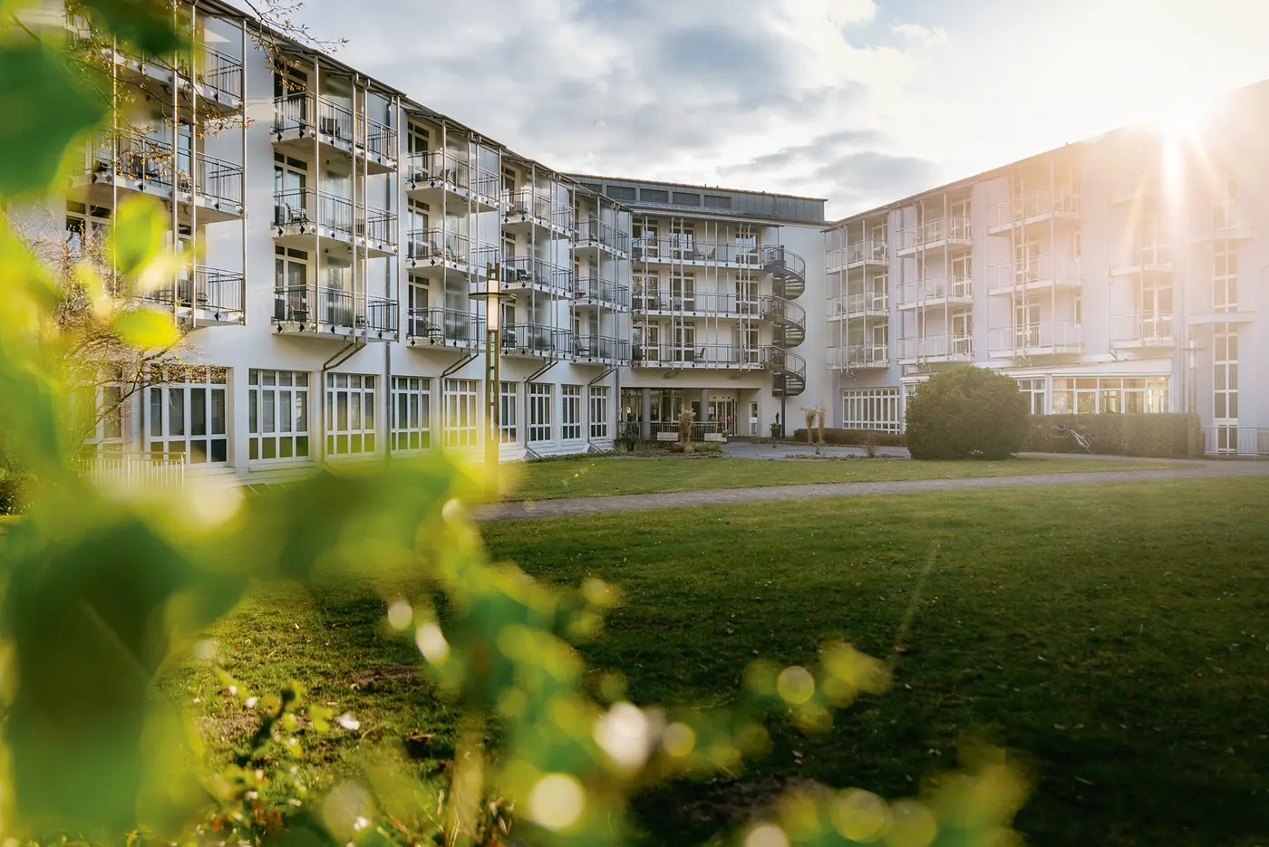 Außenansicht eines modernen, mehrstöckigen Klinikgebäudes mit vielen Balkonen. Das Gebäude ist U-förmig angelegt und von einer gepflegten Rasenfläche umgeben. Im Vordergrund sind unscharfe grüne Blätter zu sehen, im Hintergrund scheint die Sonne durch die Wolken und wirft warmes Licht auf die Fassade.