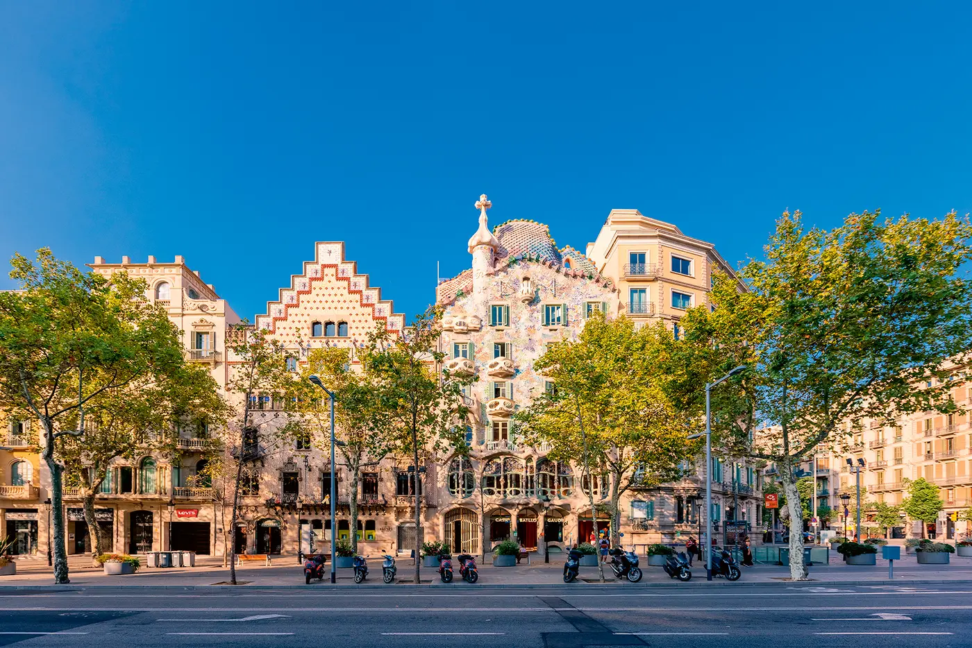 Straßenansicht in Barcelona mit der bunt verzierten Fassade eines historischen Gebäudes im modernistischen Stil, flankiert von weiteren Häusern und gesäumt von Bäumen. Im Vordergrund stehen Motorräder und Fahrräder entlang der Straße. Der Himmel ist klar und blau.
