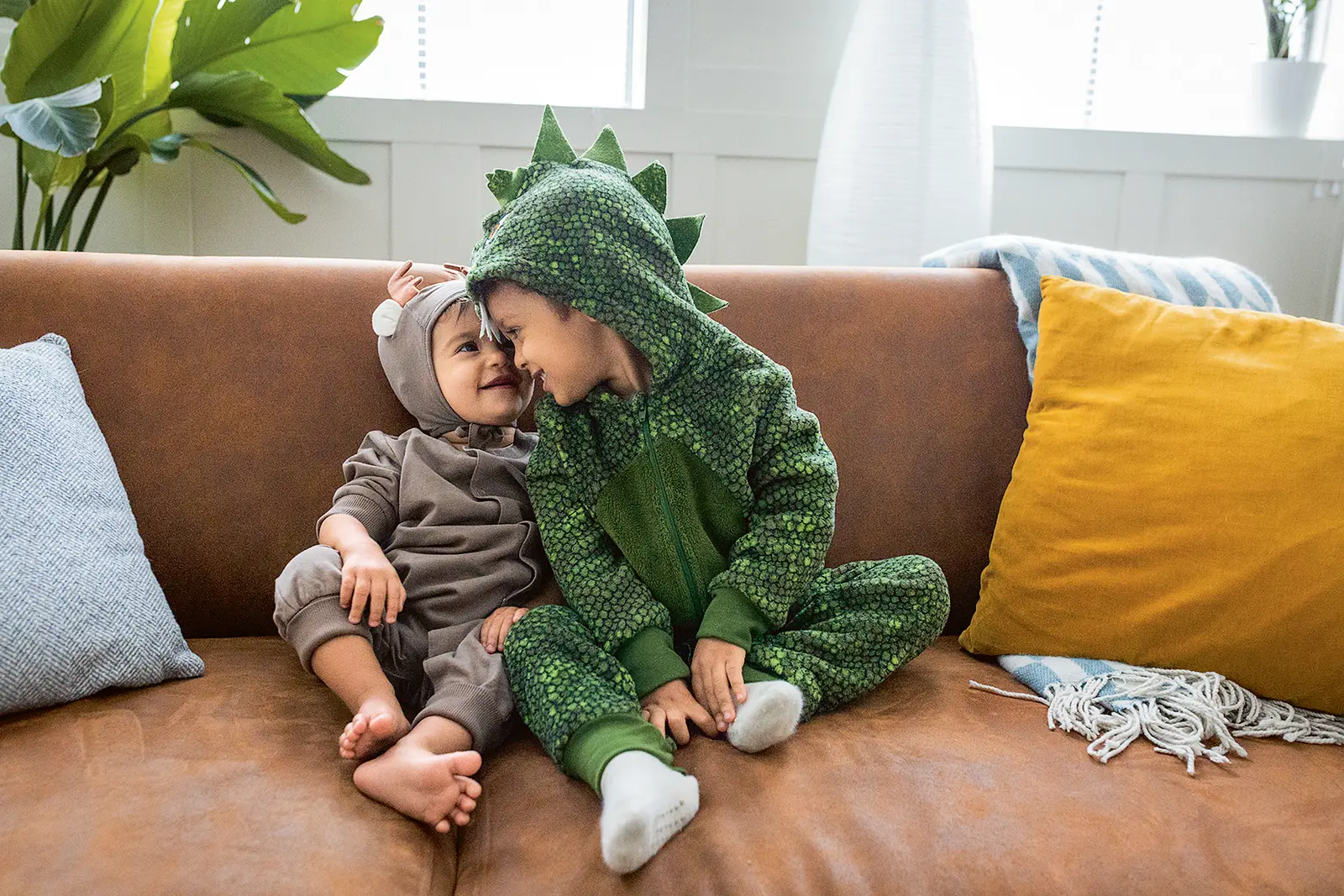 Zwei Kleinkinder sitzen verkleidet auf einem braunen Sofa und kuscheln miteinander. Das eine Kind trägt ein graues Kostüm mit Ohren, das an eine Maus erinnert, das andere ein grünes Dino-Kostüm mit Kapuze und Rückenschuppen. Sie berühren liebevoll Stirn an Stirn und lächeln sich an. Um sie herum liegen bunte Kissen und eine Decke. Im Hintergrund sind Pflanzen und ein Fenster zu sehen, durch das Tageslicht einfällt.