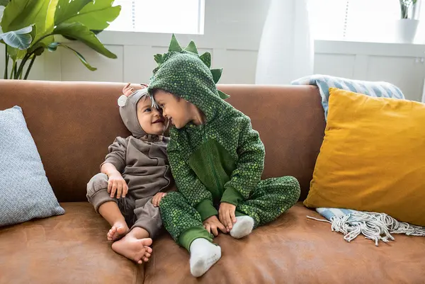Zwei Kleinkinder sitzen verkleidet auf einem braunen Sofa und kuscheln miteinander. Das eine Kind trägt ein graues Kostüm mit Ohren, das an eine Maus erinnert, das andere ein grünes Dino-Kostüm mit Kapuze und Rückenschuppen. Sie berühren liebevoll Stirn an Stirn und lächeln sich an. Um sie herum liegen bunte Kissen und eine Decke. Im Hintergrund sind Pflanzen und ein Fenster zu sehen, durch das Tageslicht einfällt.