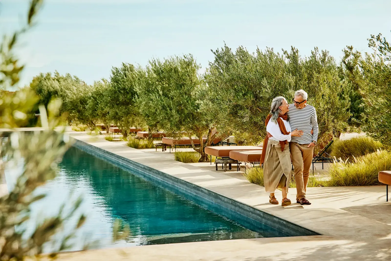 Älteres Paar spaziert Arm in Arm an einem Pool einer mediterranen Anlage mit Olivenbäumen und Sonnenliegen entlang.