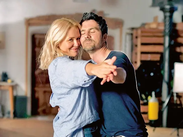 Ein Paar tanzt miteinander, während sie sich liebevoll anlächeln. Die Frau trägt ein hellblaues Hemd und der Mann ein dunkelblaues T-Shirt. Sie halten sich an den Händen, während der Mann seine Augen schließt und die Frau in die Kamera blickt. Der Hintergrund ist unscharf, mit einem rustikalen, warmen Ambiente.