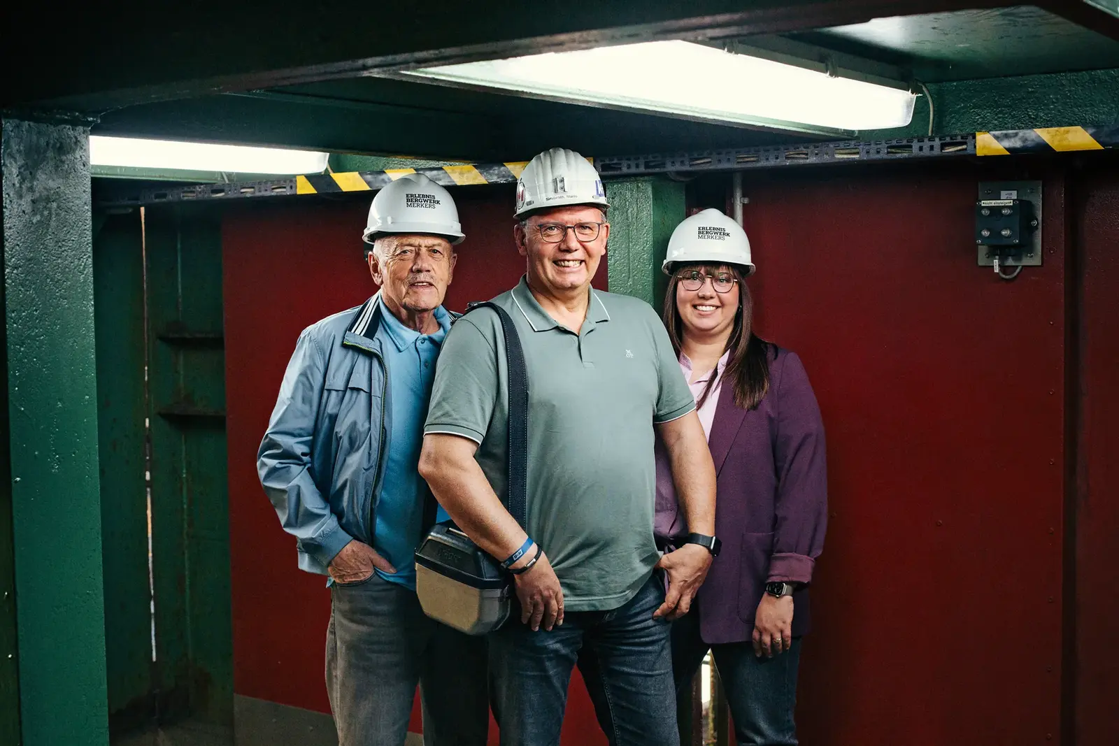Drei Personen mit Schutzhelmen stehen lächelnd in einer bergbaulichen Industrieanlage.
