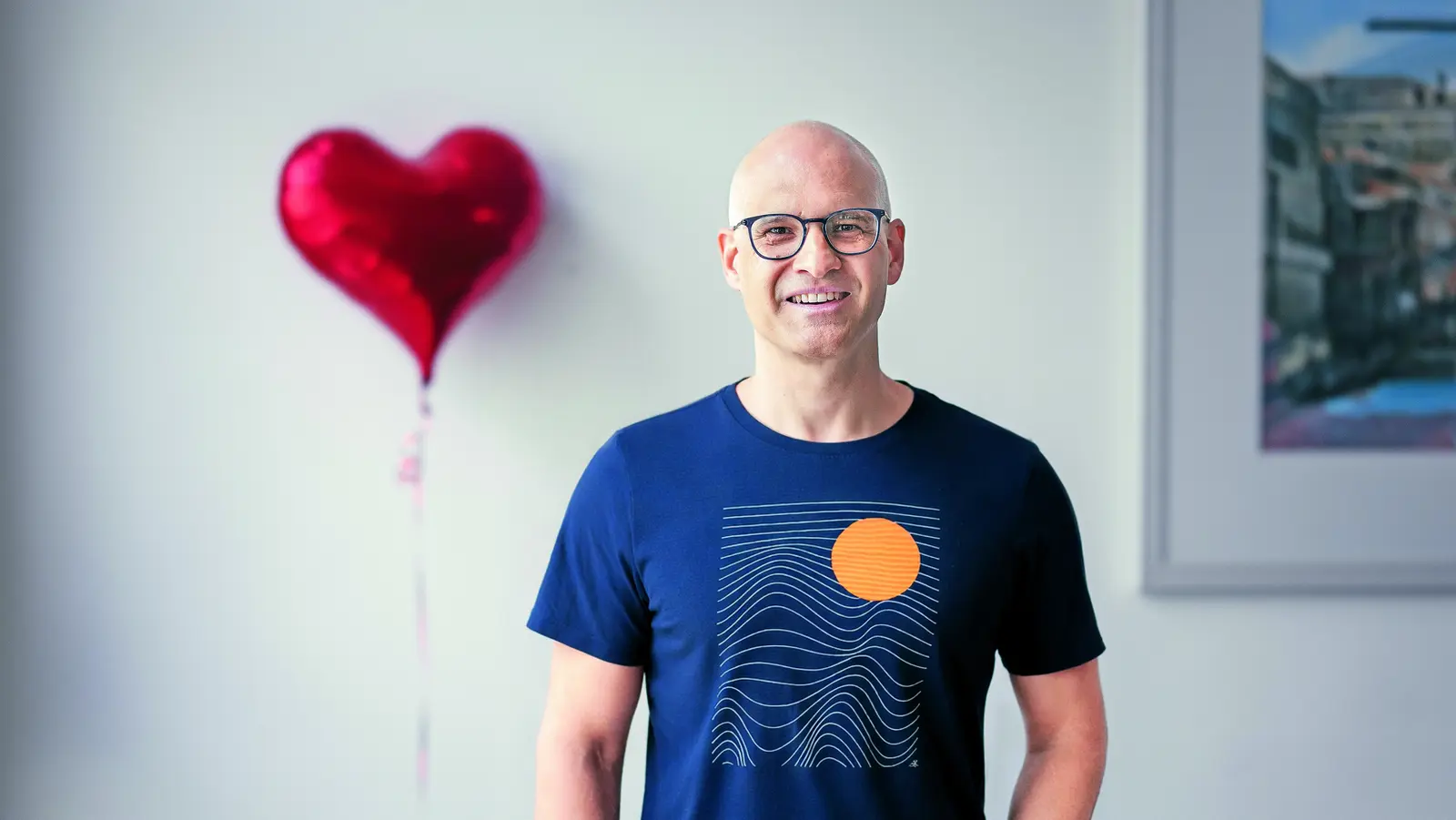 Das Bild zeigt einen Mann mit Glatze und Brille der in die Kameralächelt. Im Hintergrund ich ein roter Folienluftballon in der Form eines Herzes zu sehen. 