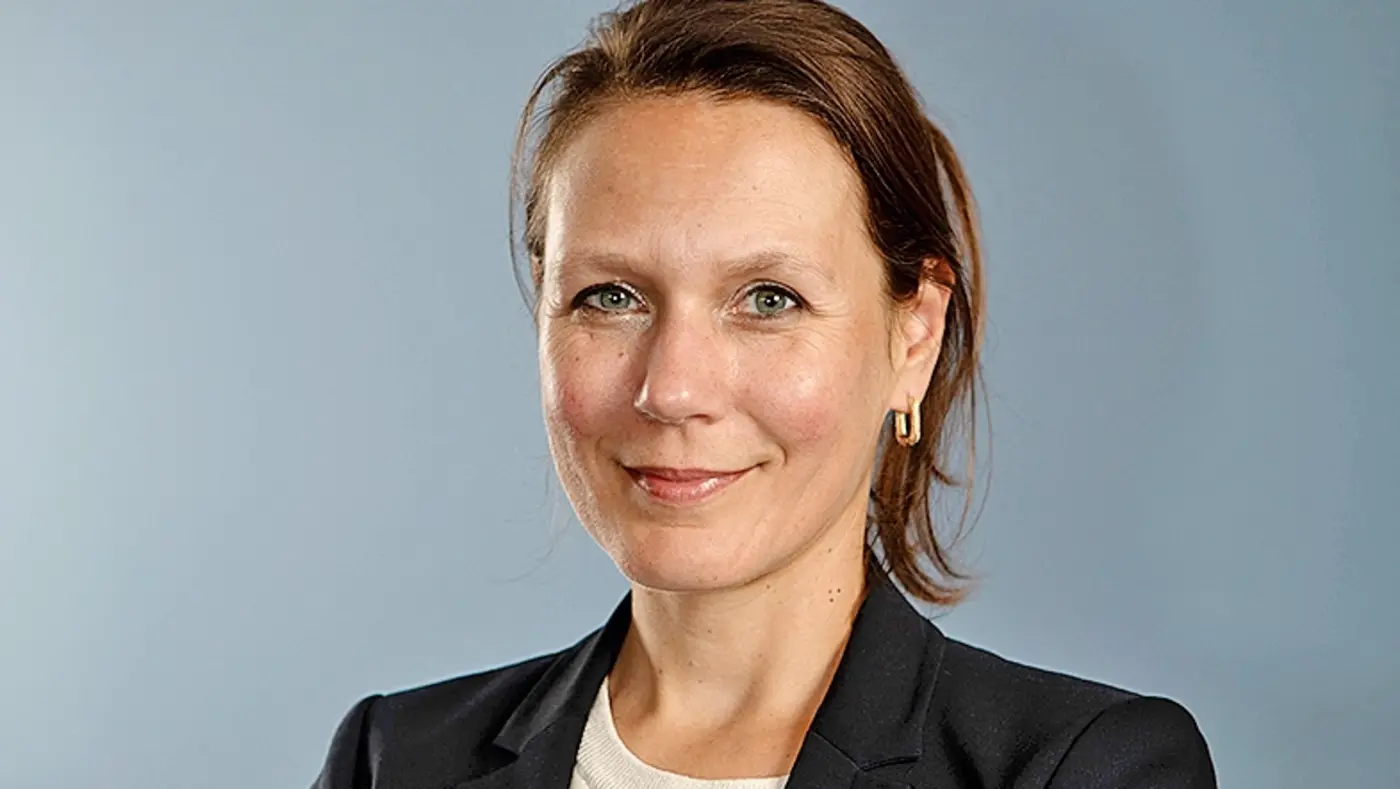 Katja Wolter lächelt mit verschränkten Armen im dunklen Sakko vor hellblauem Hintergrund.