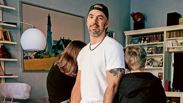 Ein Mann steht lässig an eine graue Couch gelehnt, trägt eine weiße T-Shirt, hellblaue Jeans, weiße Sneakers und eine nach hinten getragene Basecap. Er lächelt in die Kamera. Zwei Kinder sitzen mit dem Rücken zur Kamera auf der Couch – eines lehnt sich nach vorne, das andere schaut seitlich weg. Im Hintergrund sind ein Regal mit Büchern und Spielen, ein großes Wandbild mit Leuchtturm-Motiv und eine moderne Stehlampe zu sehen.