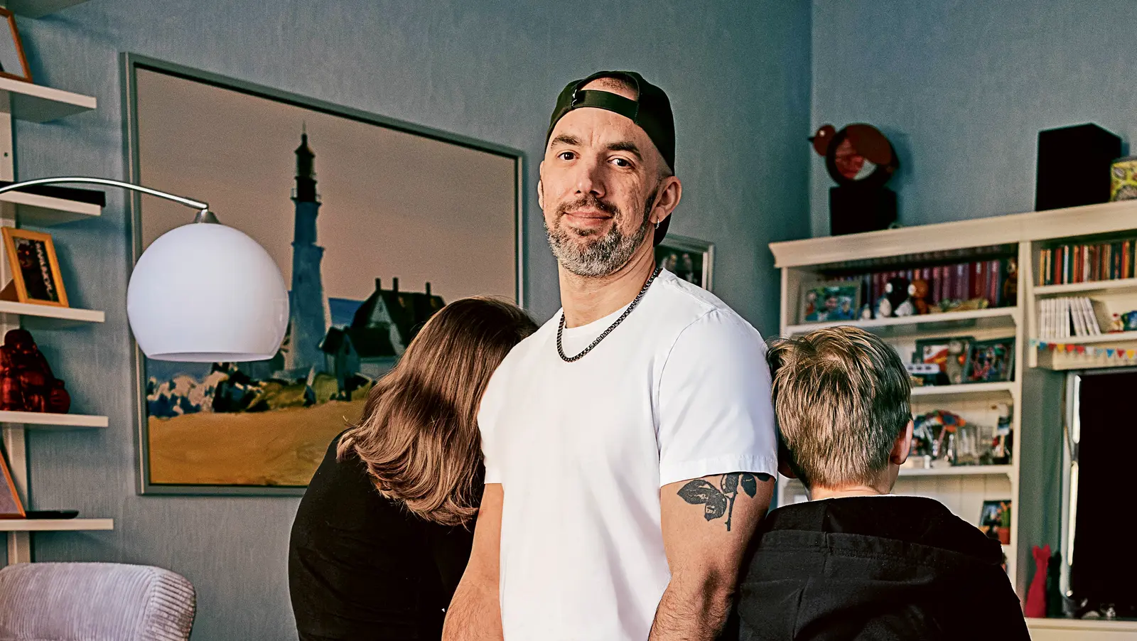 Ein Mann steht lässig an eine graue Couch gelehnt, trägt eine weiße T-Shirt, hellblaue Jeans, weiße Sneakers und eine nach hinten getragene Basecap. Er lächelt in die Kamera. Zwei Kinder sitzen mit dem Rücken zur Kamera auf der Couch – eines lehnt sich nach vorne, das andere schaut seitlich weg. Im Hintergrund sind ein Regal mit Büchern und Spielen, ein großes Wandbild mit Leuchtturm-Motiv und eine moderne Stehlampe zu sehen.