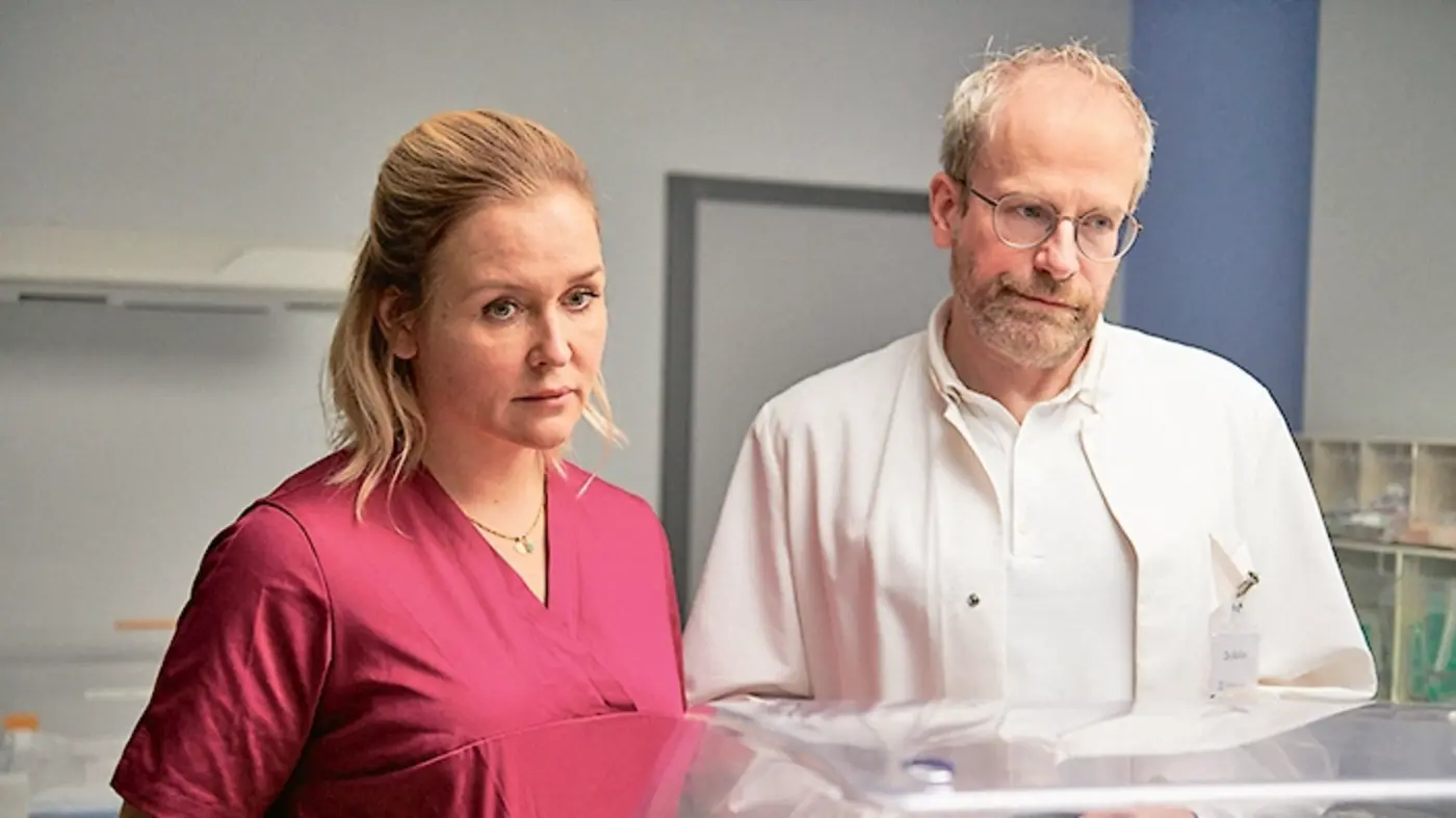 Ein Arzt und eine Hebamme stehen nebeneinander und blicken aufmerksam auf einen Inkubator. Die Hebamme trägt ein pinkes Hemd, der Arzt einen weißen Kittel. Beide haben einen ernsten Gesichtsausdruck, während sie die Situation beobachten.