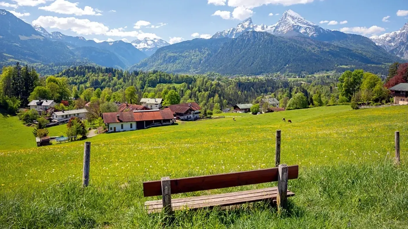Bank auf einer grünen Wiese mit Blick auf ein Dorf und dahinter Berge unter blauem Himmel mit Wolken