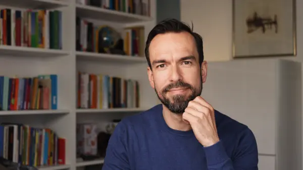 Ein Mann mit Bart sitzt nachdenklich vor einem Bücherregal und blickt in die Kamera, eine Hand am Kinn.