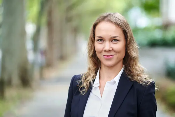 Porträtaufnahme einer Frau im Business-Outfit (dunkles Sakko, weiße Bluse), die freundlich in die Kamera blickt. Im Hintergrund ist eine verschwommene Allee erkennbar.