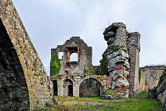 Ruinen einer alten Burg, von der nur noch zerfallene Mauern und Bögen übrig sind. Efeu und Pflanzen haben sich an den Steinen festgesetzt.