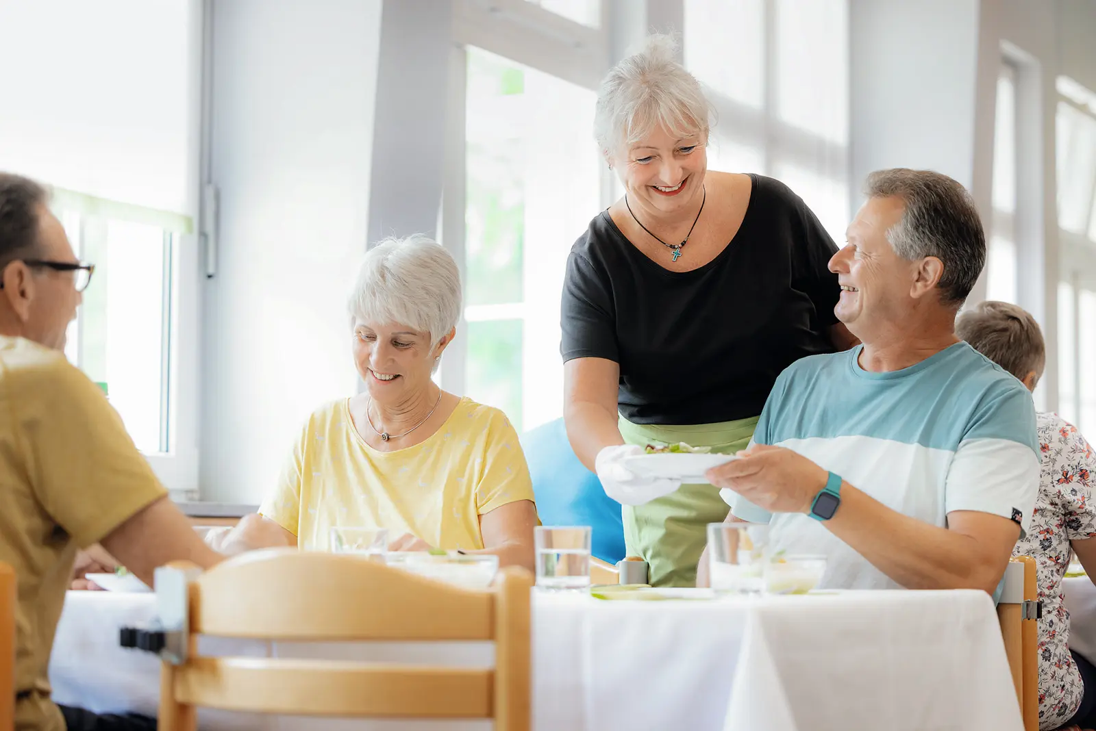 In einem hellen Speisesaal reicht eine Mitarbeiterin einem lächelnden Mann einen Teller; weitere ältere Gäste sitzen am Tisch und unterhalten sich — eine freundliche, gemeinschaftliche Szene in der Reha.