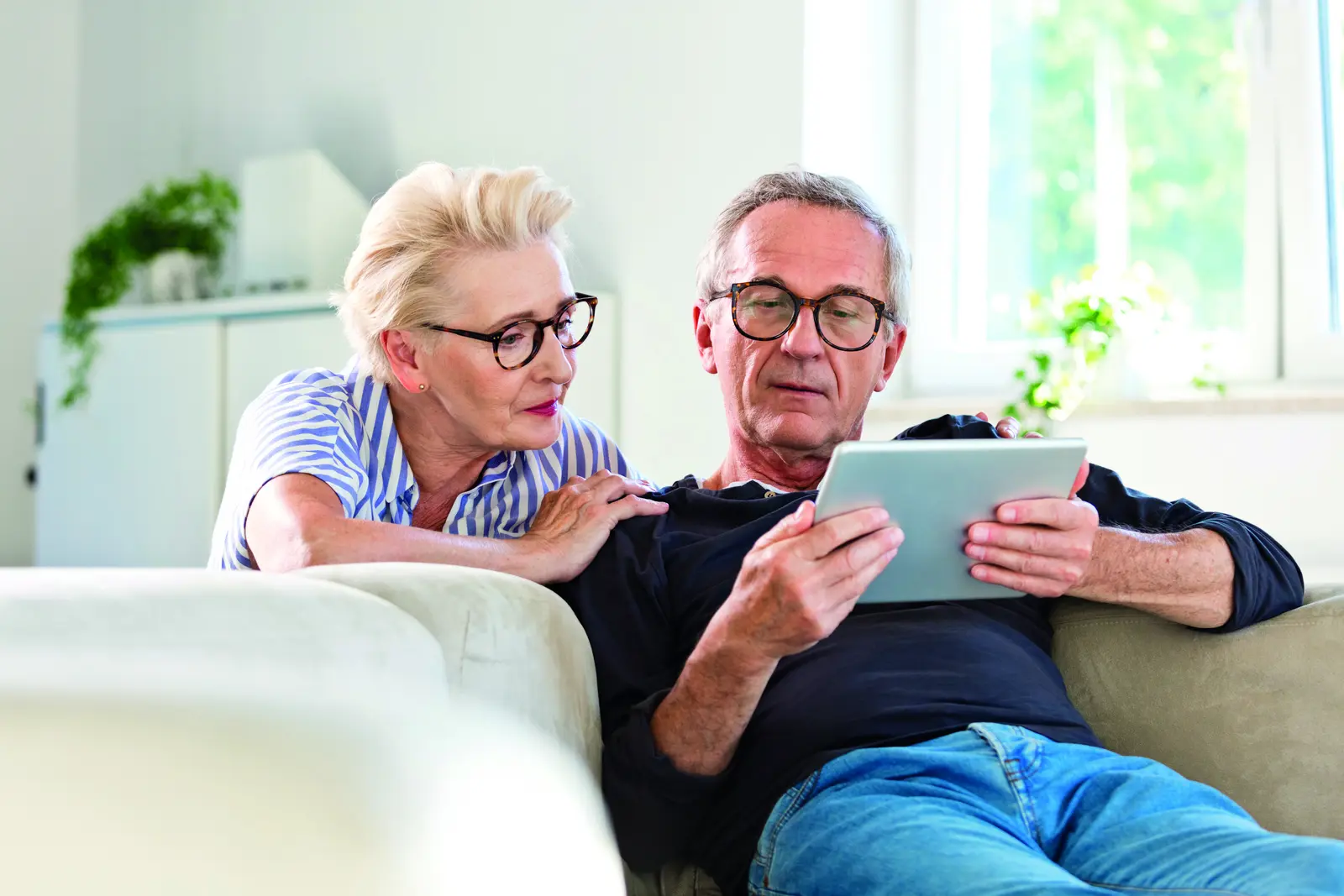 Ein älteres Paar sitzt gemeinsam auf dem Sofa und schaut auf ein Tablet. Die Frau legt ihre Hand auf die Schulter des Mannes, beide tragen Brillen und wirken konzentriert auf den Bildschirm.