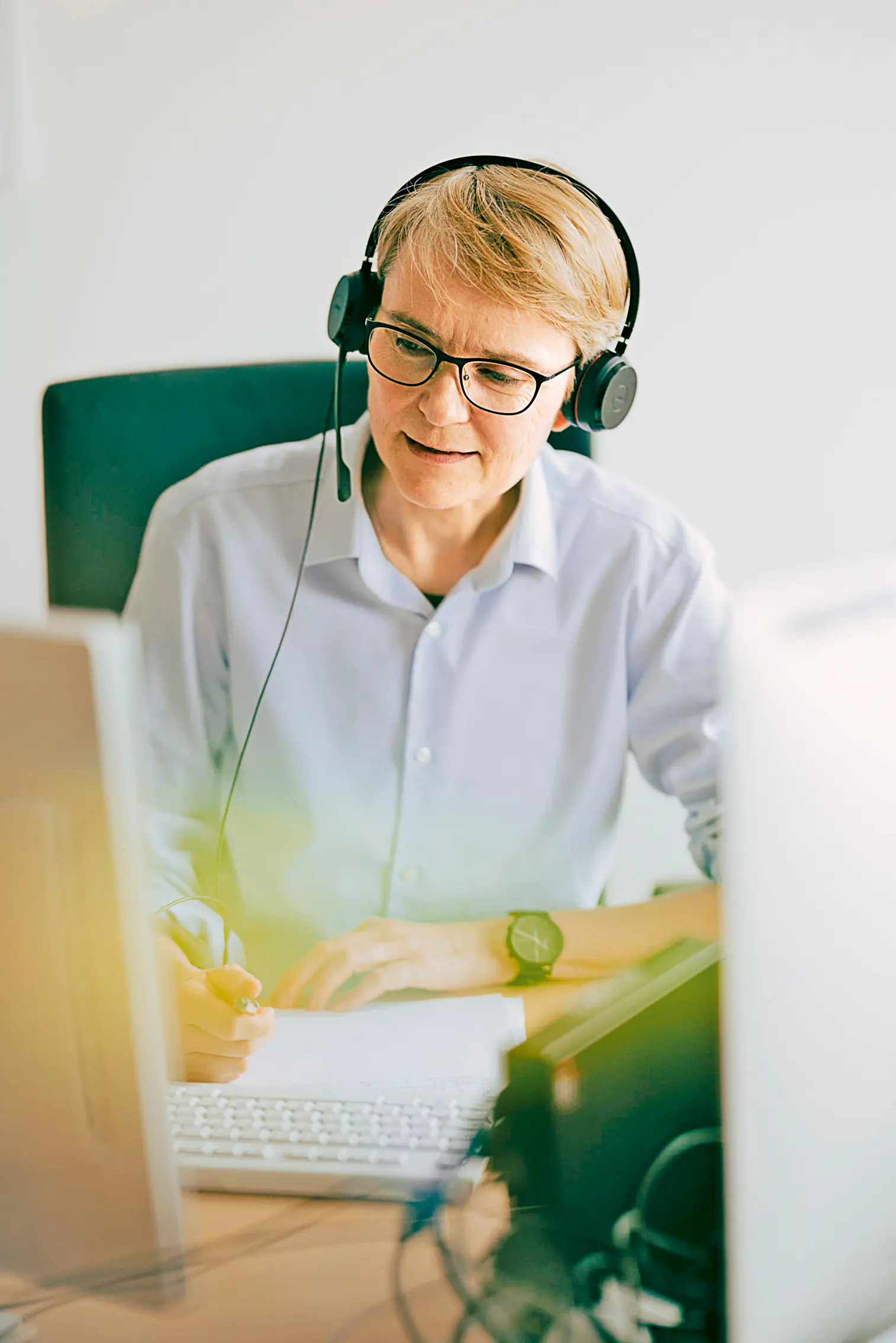 Eine Person mit kurzen blonden Haaren sitzt am Schreibtisch, trägt ein Headset und schaut auf den Bildschirm. Sie schreibt etwas auf und wirkt konzentriert bei der Arbeit.