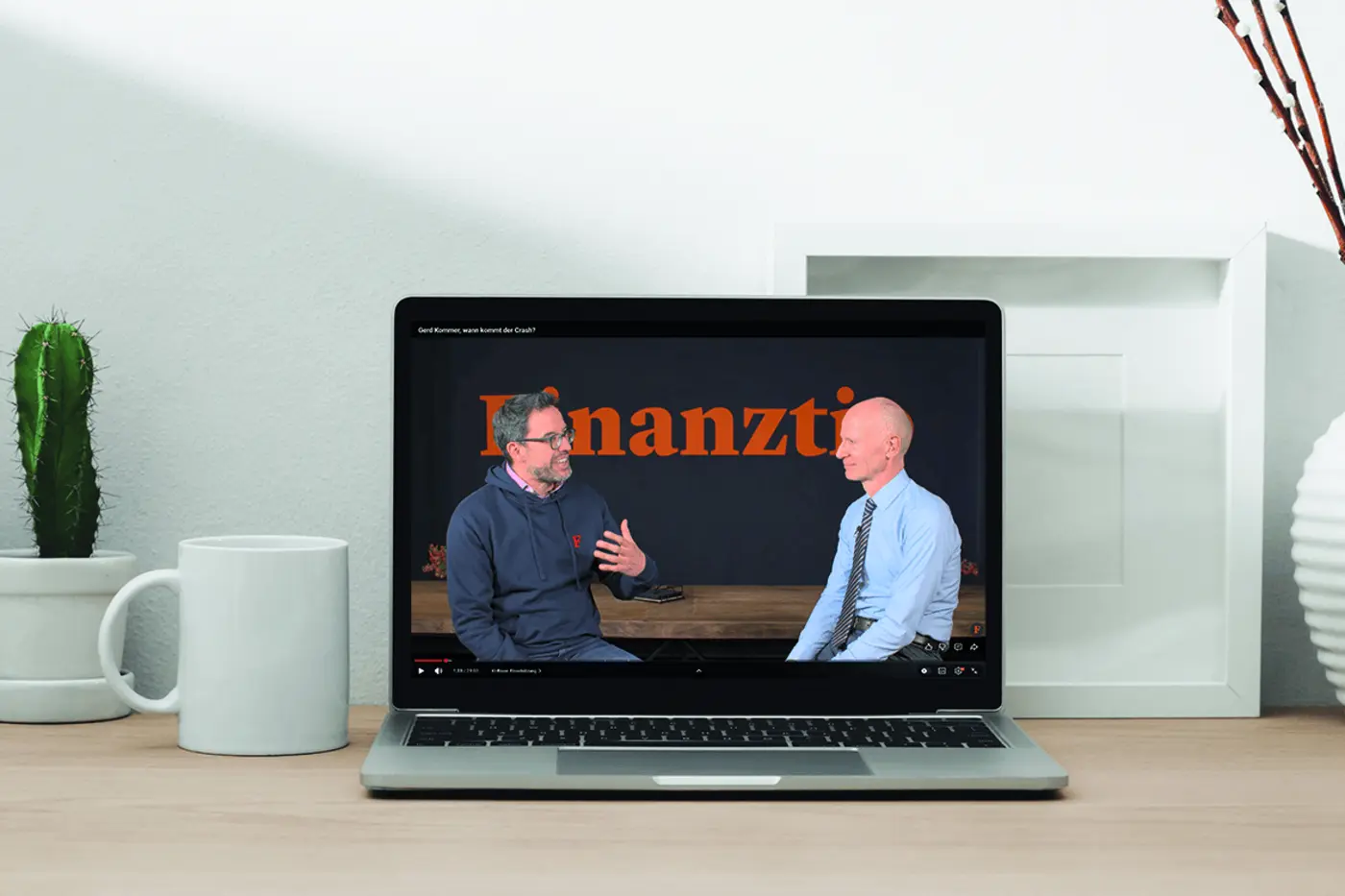 Ein auf einem Holzschreibtisch stehender Laptop, auf dessen Bildschirm ein Interview-Video von „Finanztip“ mit zwei Männern zu sehen ist, daneben eine Tasse und ein kleiner Kaktus.