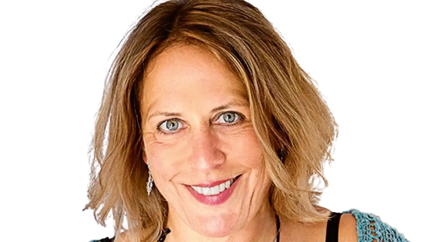 Porträt einer lächelnden Frau mit schulterlangem, blondem Haar. Sie trägt ein schwarzes Top, eine blaue Strickjacke und eine Halskette.