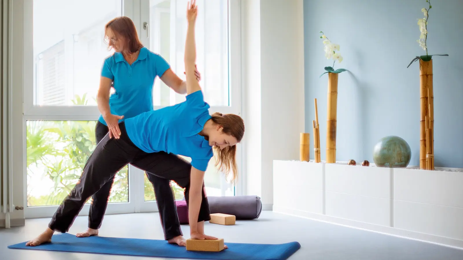 Eine Frau führt auf einer Yogamatte Übungen aus, während eine andere Frau ihr assistiert.
