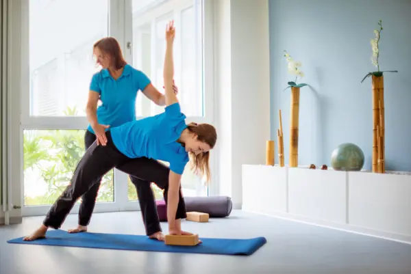 Eine Frau führt auf einer Yogamatte Übungen aus, während eine andere Frau ihr assistiert.
